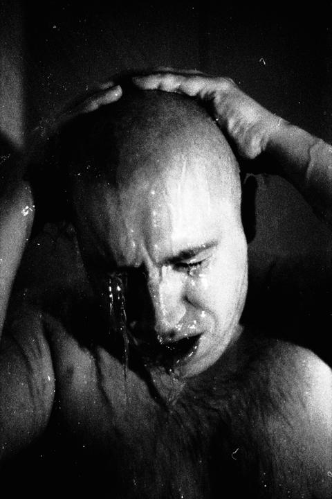 A man with a shaved head holds his head as water pours over his face and body.