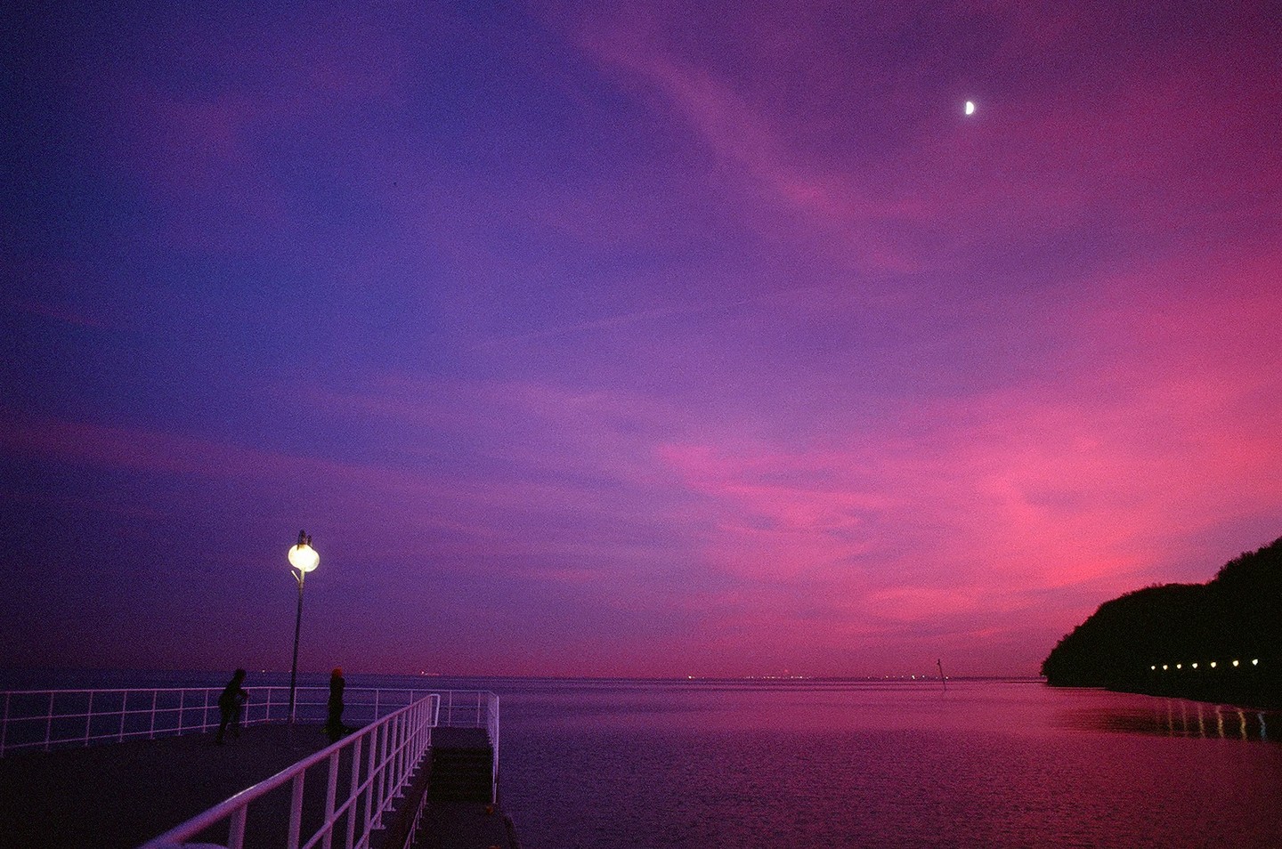Two people stand on a pier under a vibrant pink and purple sunset sky with a crescent moon.