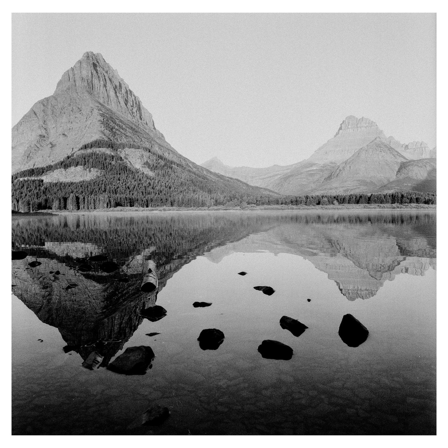 A mountain range with a forest at its base is reflected in a calm lake with rocks in the foreground.