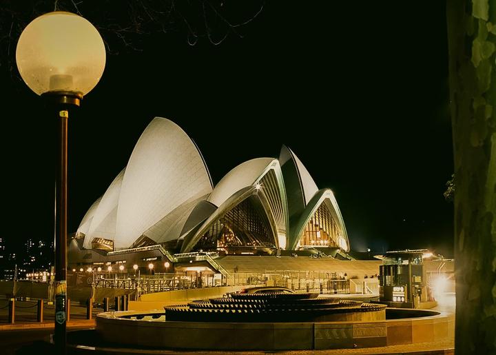 The Sydney Opera House is illuminated at night, with a street light and a fountain in the foreground.