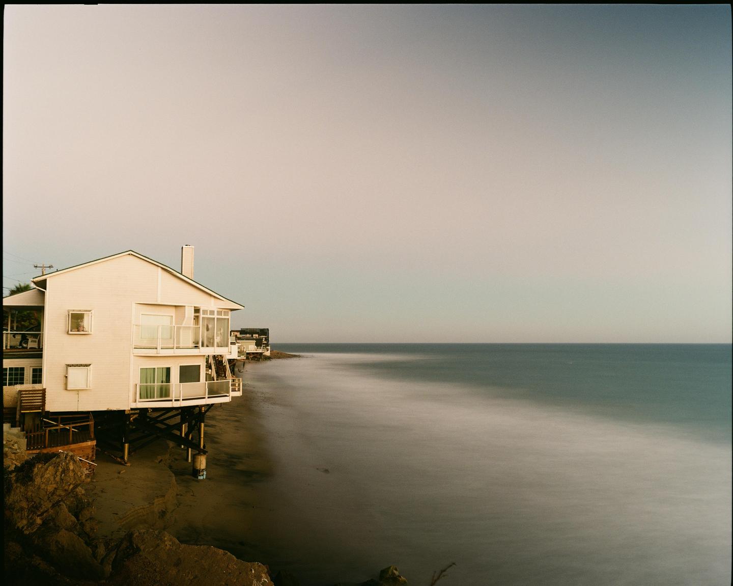 A house on stilts stands on a sandy beach next to the ocean under a clear sky.