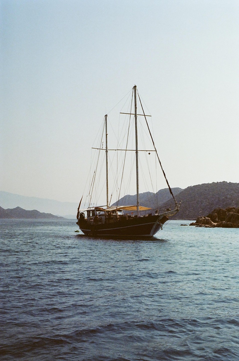 A sailboat with two masts floats on the water near a rocky shore with mountains in the background.
