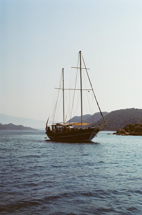 A sailboat with two masts floats on the water near a rocky shore with mountains in the background.