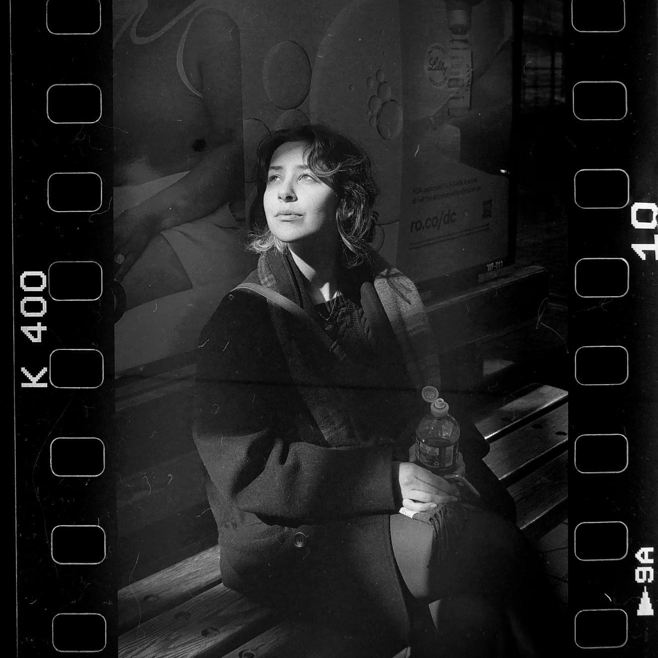 A woman sits on a bench, holding a bottle, and looking up.