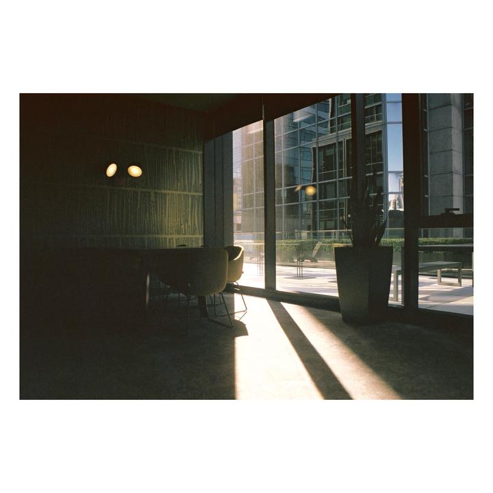Sunlight streams through a large window into a room with chairs and a plant, revealing a modern building outside.