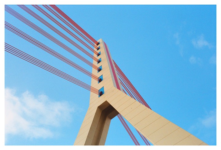 A bridge support with red cables extends into a clear blue sky with a few clouds.
