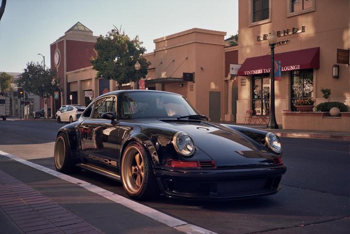A black Porsche with gold wheels is parked on a street in front of buildings with awnings.