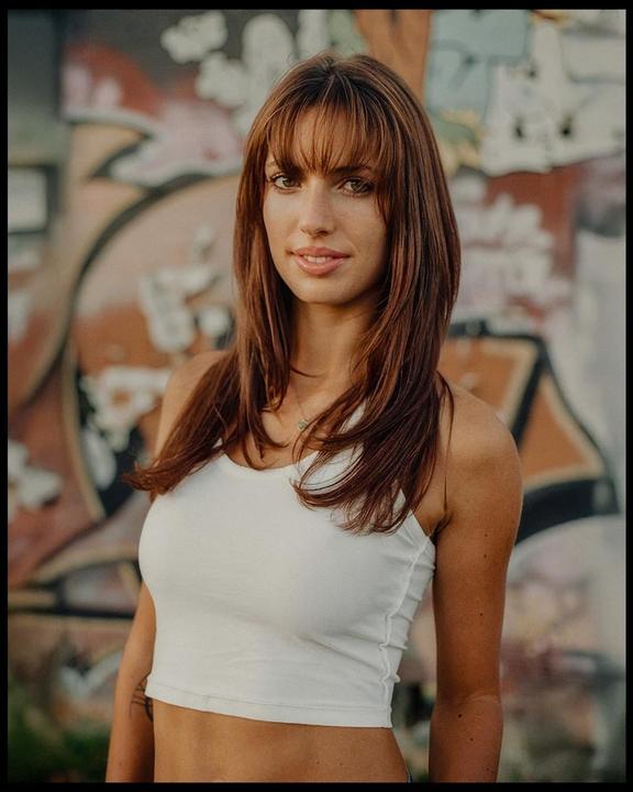 A woman with long hair and bangs wears a white crop top and stands in front of a graffiti-covered wall.