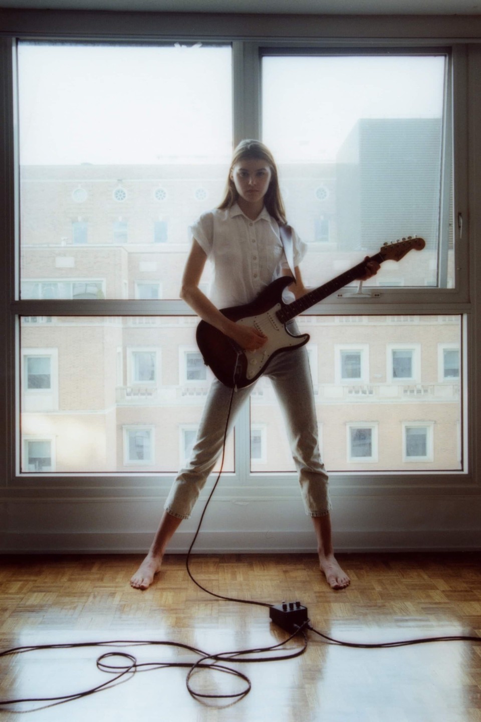 A woman stands barefoot in front of a window, playing an electric guitar connected to a pedal on a wooden floor.