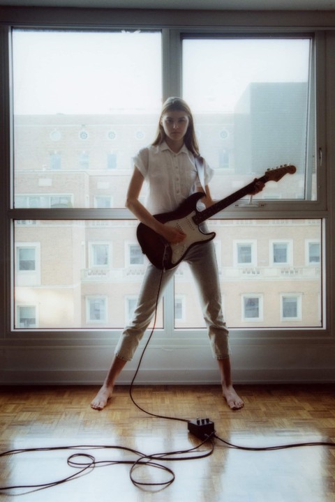 A woman stands barefoot in front of a window, playing an electric guitar connected to a pedal on a wooden floor.