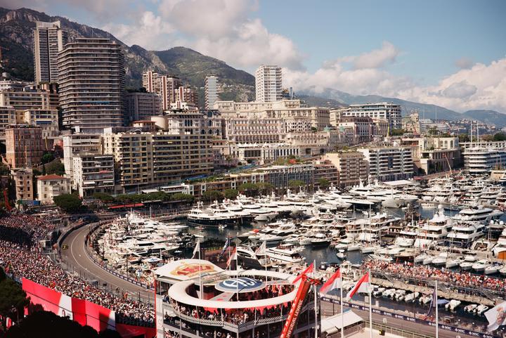 A large crowd of people watches a race car on a track next to a harbor filled with yachts, with a city and mountains in the background.