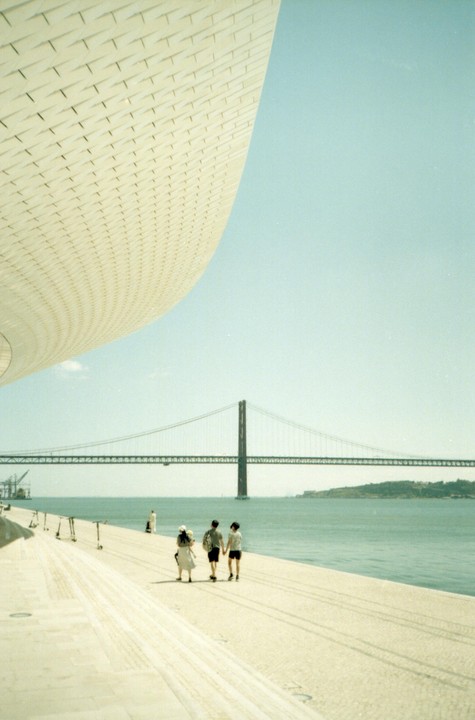 Three people walk along a waterfront promenade with a suspension bridge in the background and a modern building curving into the sky.