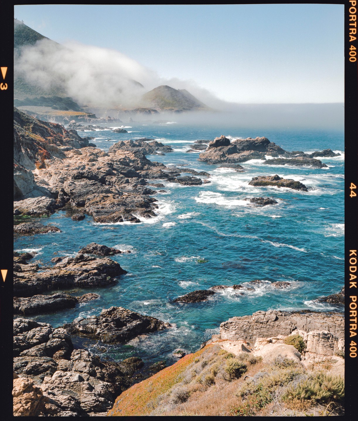 A rocky coastline with blue water and a hazy mountain in the background.