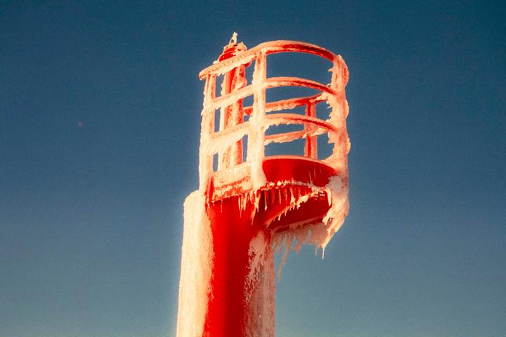 A red lighthouse is covered in ice and icicles against a clear blue sky.