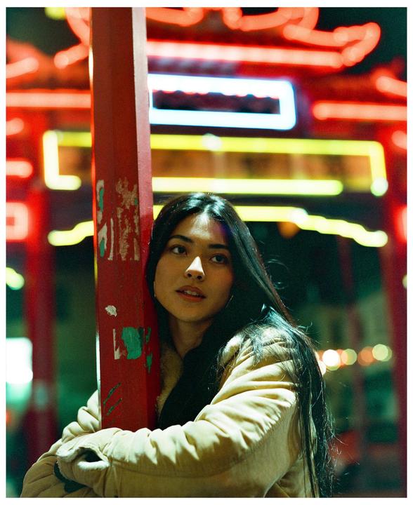 A woman with long dark hair leans against a red pillar with colorful neon lights in the background at night.