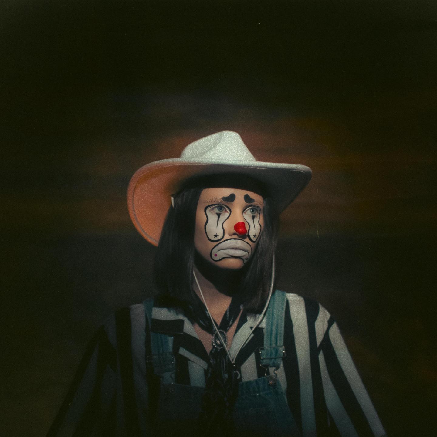 A woman wearing a cowboy hat and clown makeup looks up and to the right.