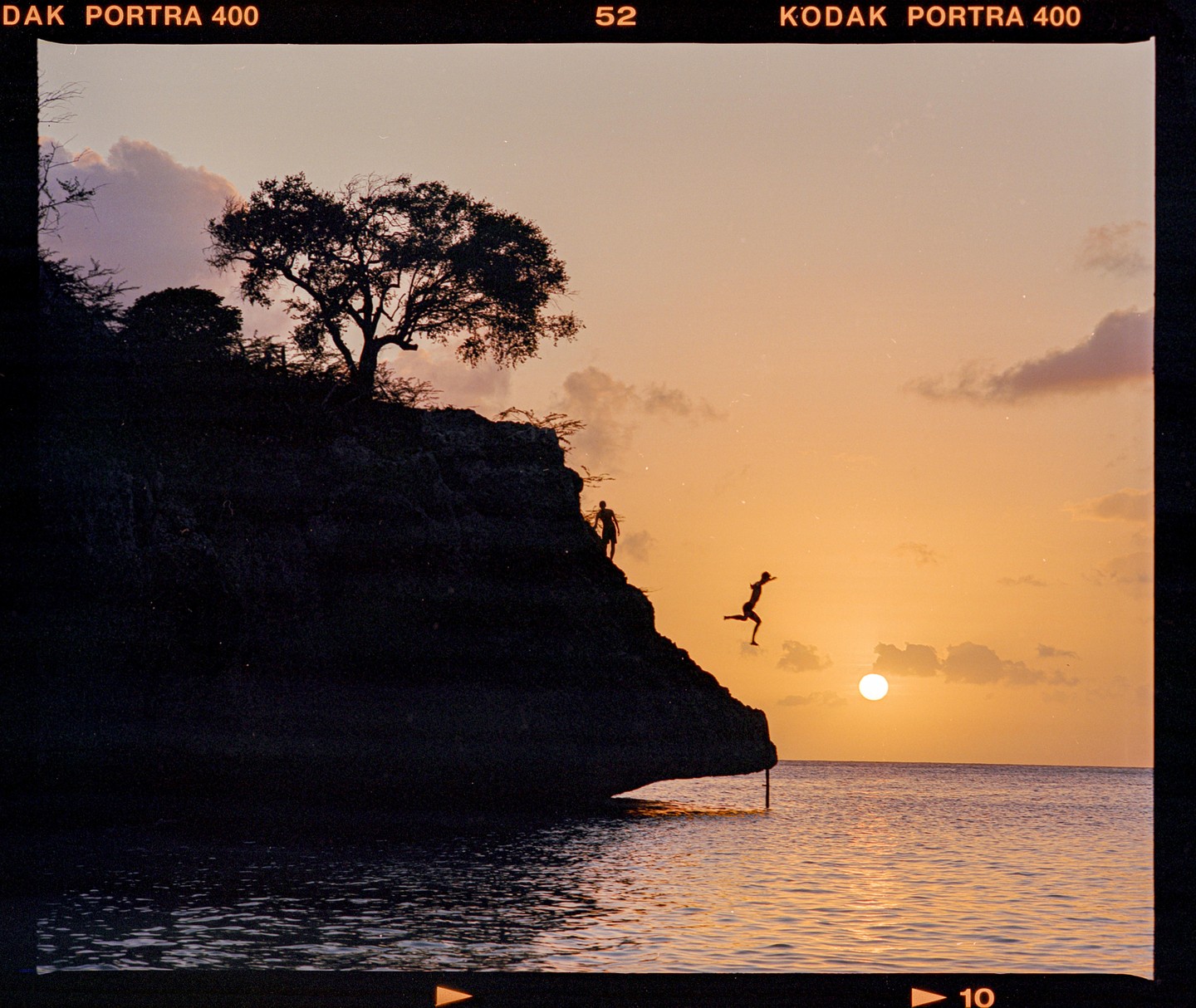 A person jumps from a cliff into the ocean at sunset while another person stands on the cliff.