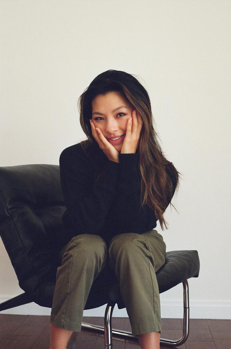 A woman with long hair sits in a chair, smiling with her hands on her cheeks.