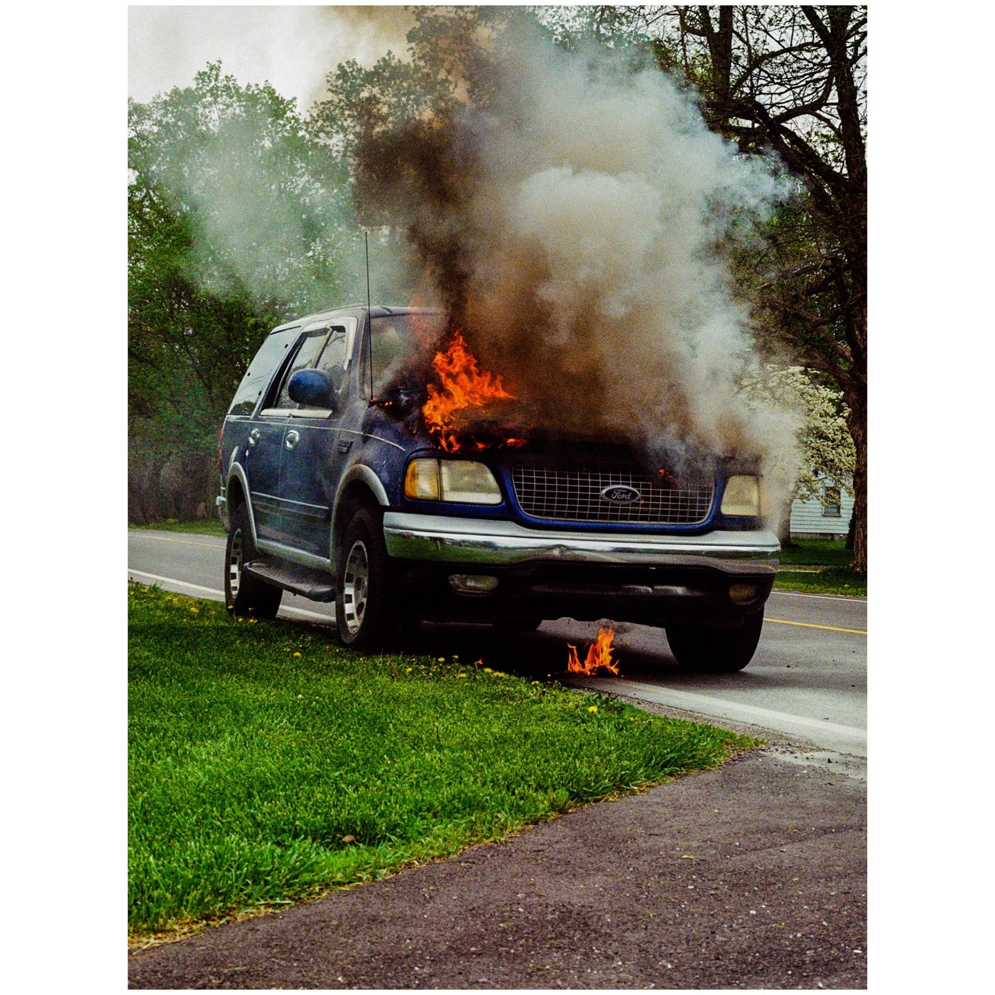 A blue SUV is engulfed in flames and smoke on the side of a road.