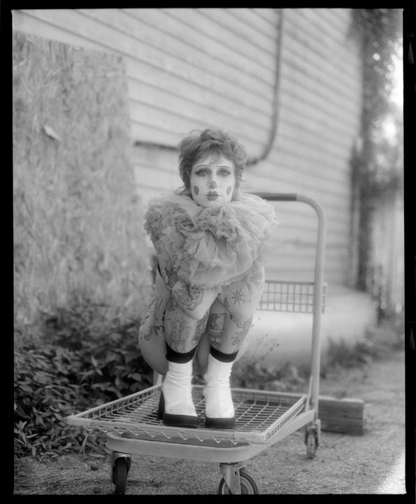 A person in clown makeup and a ruffled collar crouches on a cart.