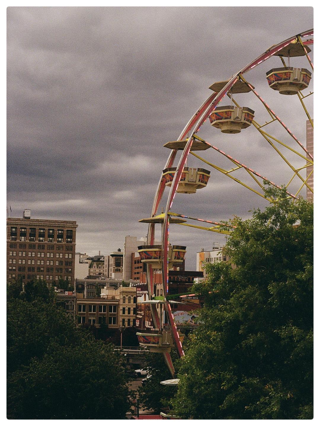 A ferris wheel stands tall against a cloudy sky, with city buildings and trees in the background.