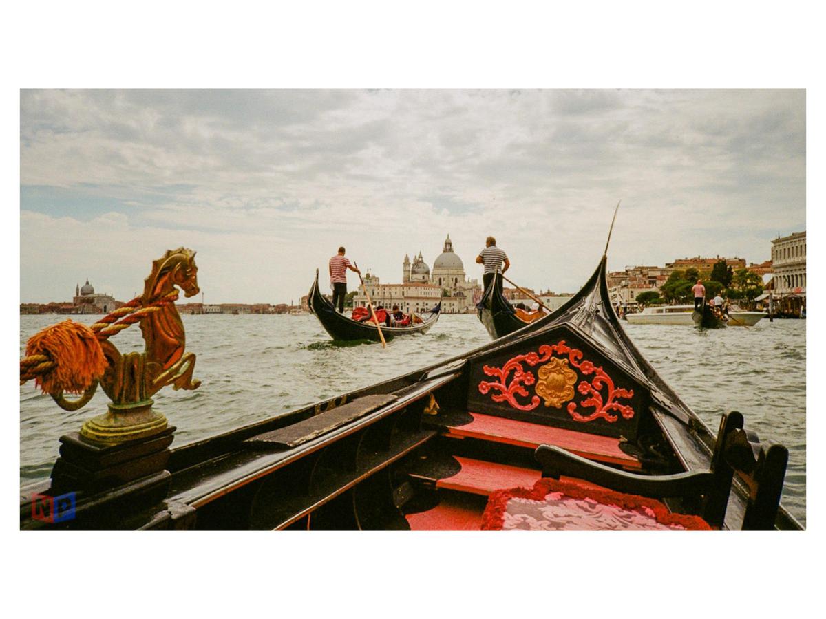 A gondola with a seahorse ornament in the foreground is on a canal with other gondolas and buildings in the background.