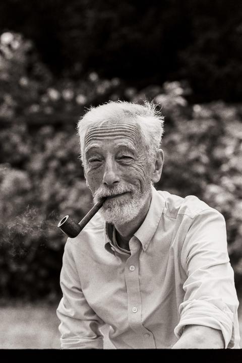 A man with a white beard and hair smiles while smoking a pipe.