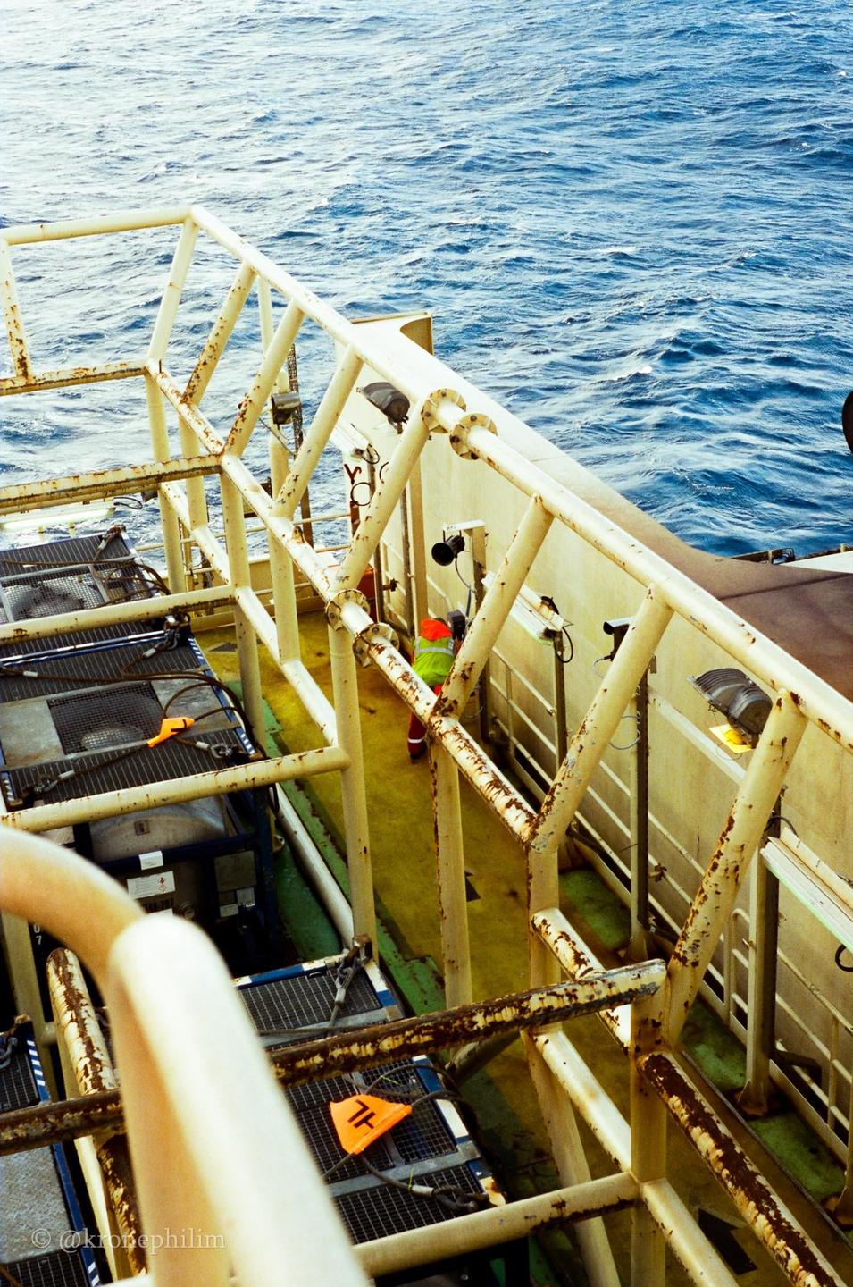A person in a safety vest walks along a narrow walkway on an offshore platform next to the ocean.