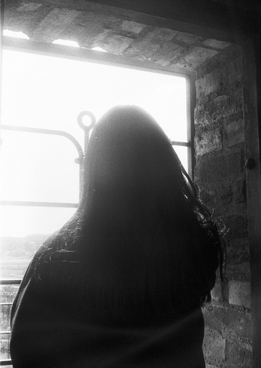 A person with long hair looks out a window with a bright light outside.