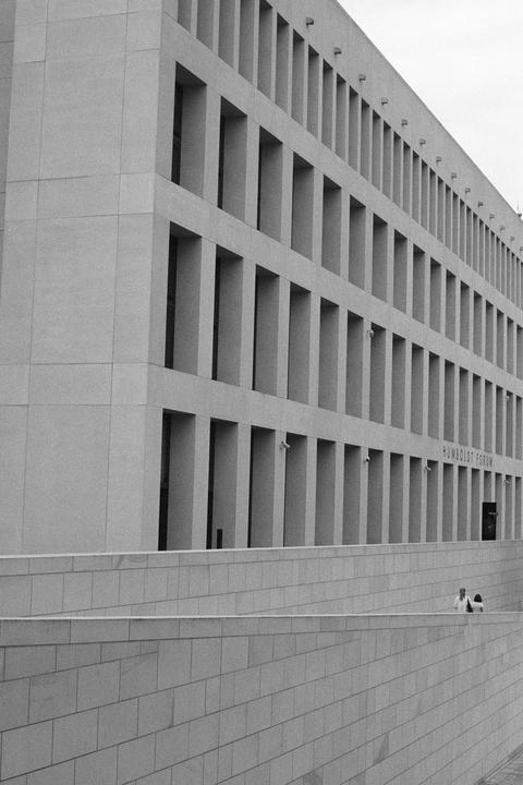 A large building with many rectangular windows is seen from an angle, with two people visible behind a wall in the lower right.