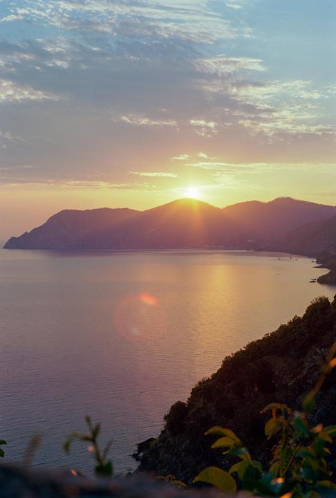 The sun sets over a body of water with mountains in the background and foliage in the foreground.