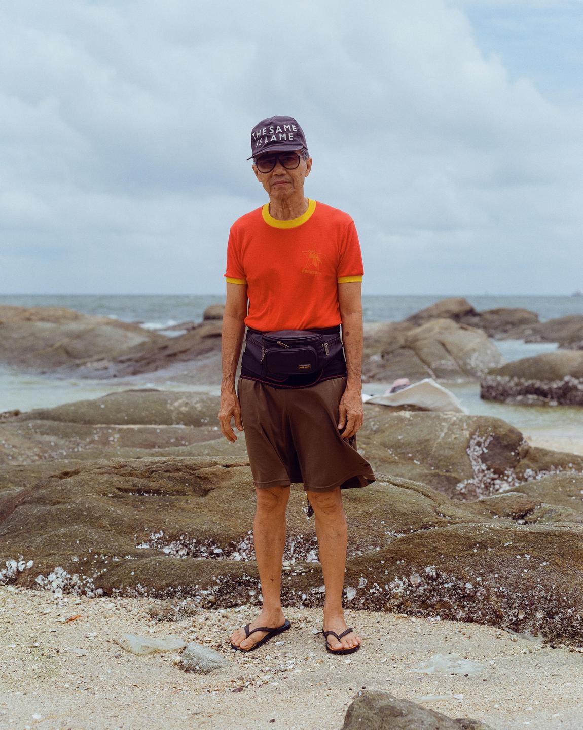 A man wearing a cap, sunglasses, and a fanny pack stands on a rocky beach.