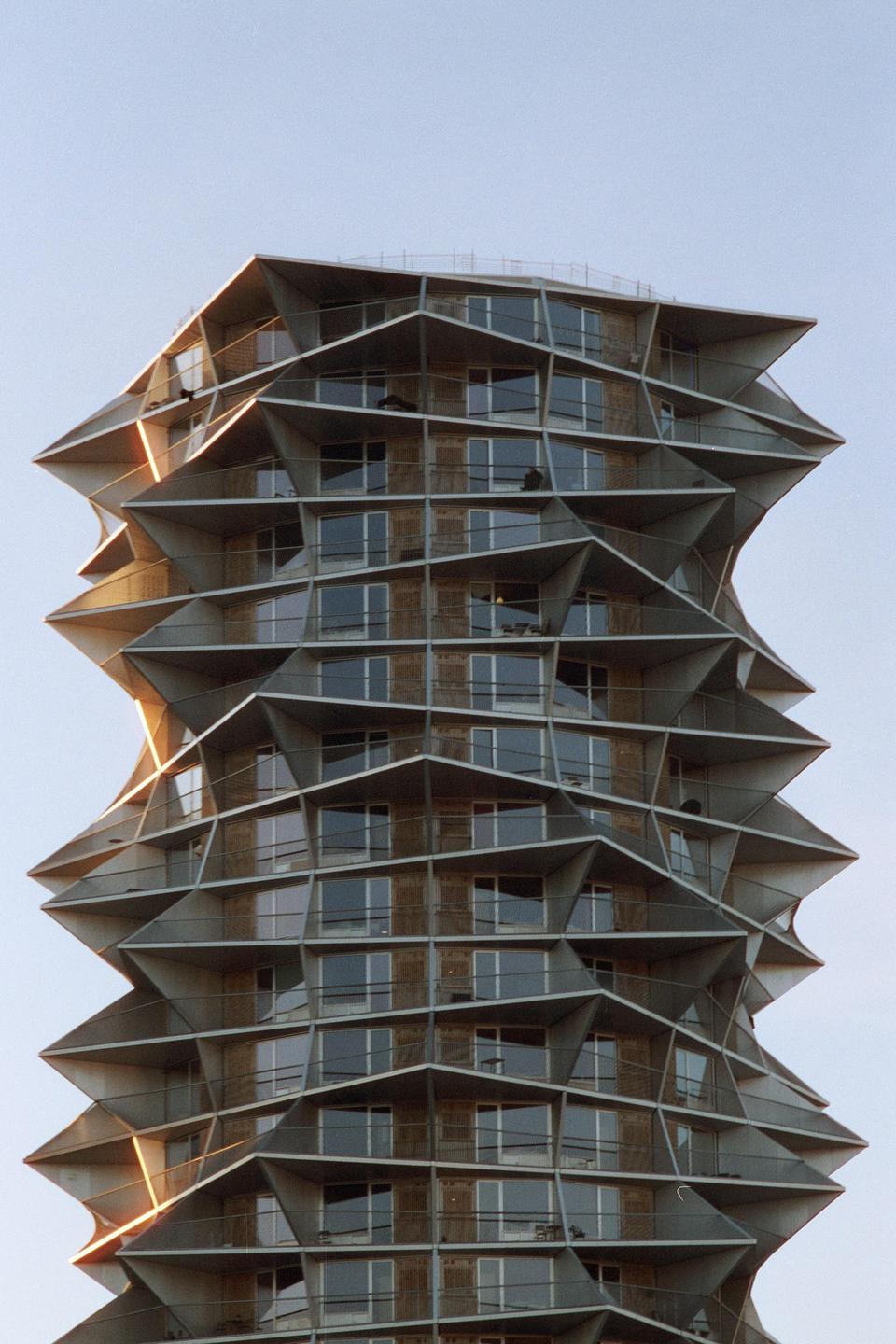 A tall, modern building with a unique, undulating facade and many windows is shown against a clear sky.