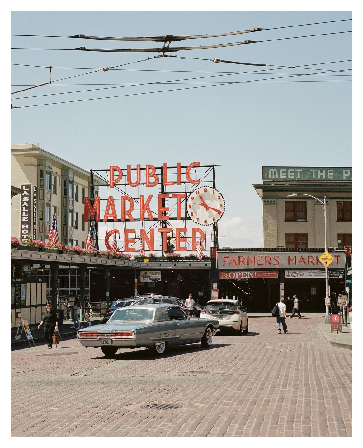 A vintage car drives on a brick street in front of the Public Market Center sign and Farmers Market entrance.