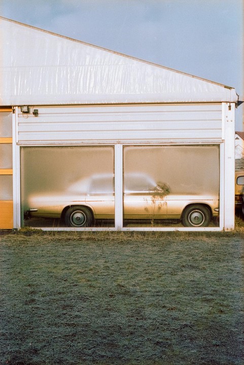 A gold car is parked inside a garage with frosted windows, with a patch of grass in the foreground.
