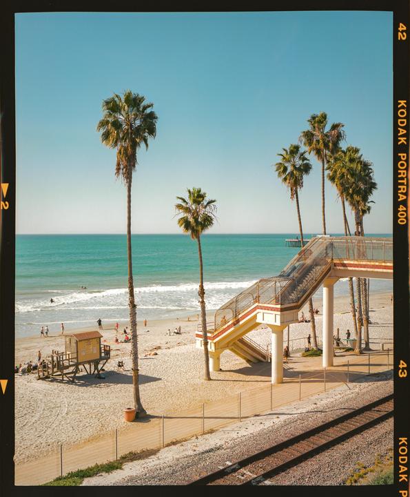 A beach with palm trees, a lifeguard stand, and a pedestrian bridge over railroad tracks.