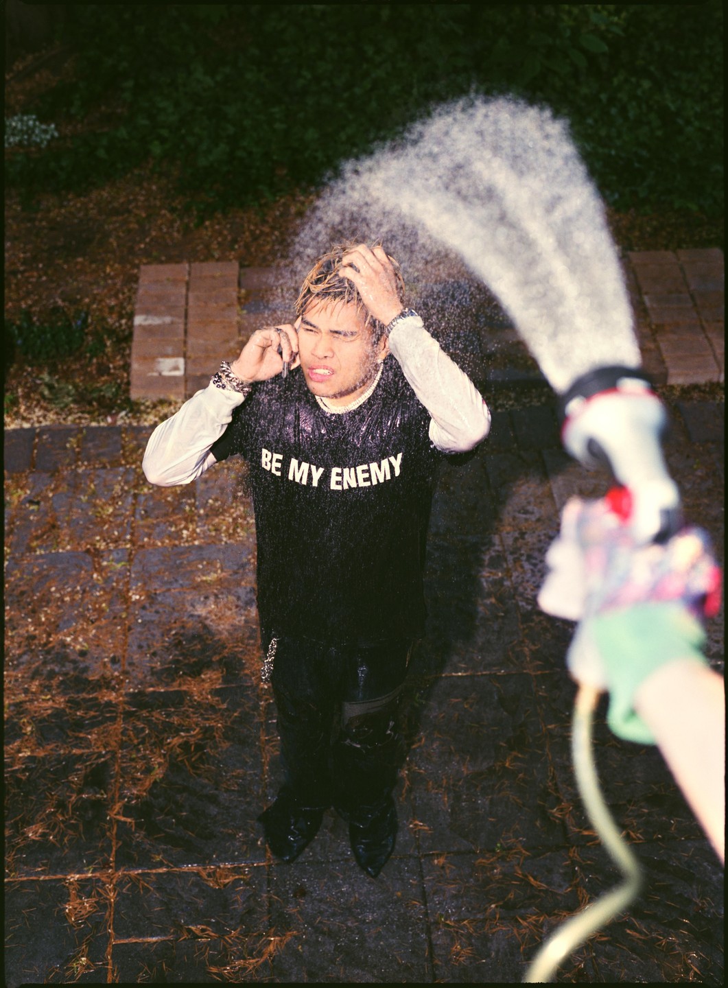 A man with bleached hair is being sprayed with water from a hose, looking up with his hands in his hair.