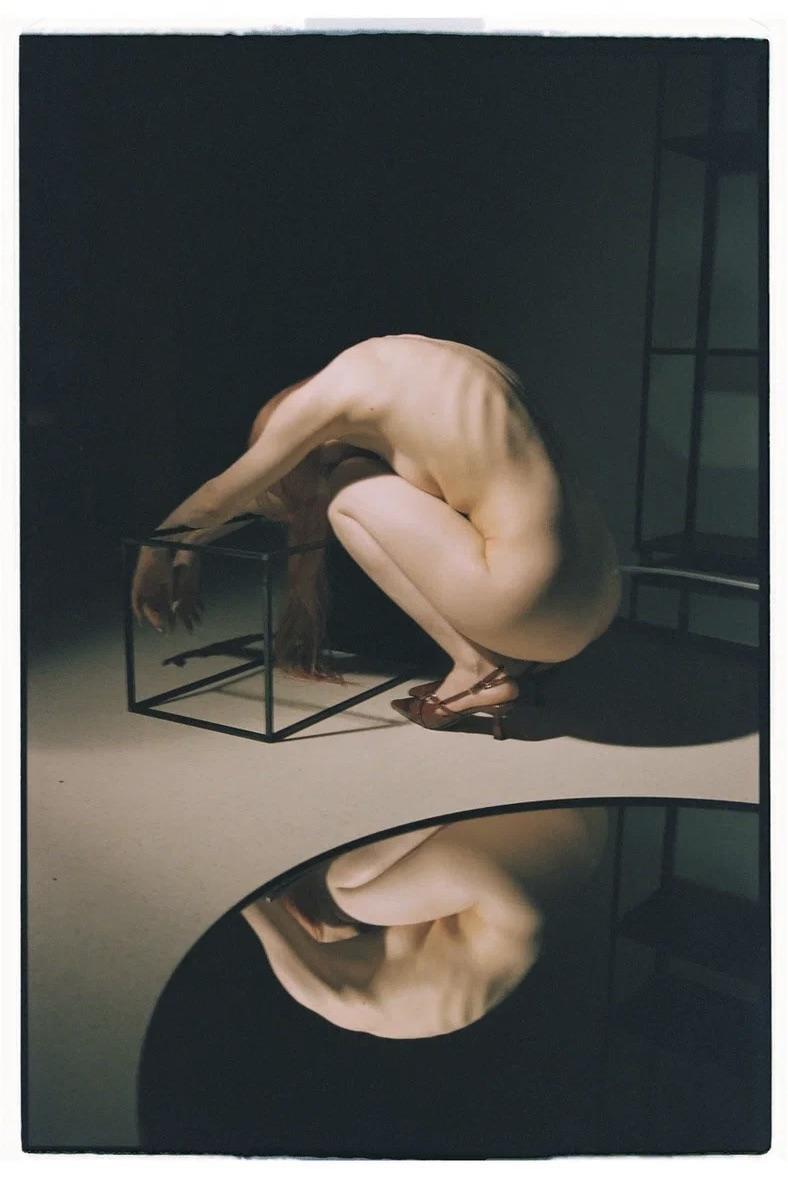 A nude woman crouches over a wire cube, with her reflection visible in a circular mirror on the floor.