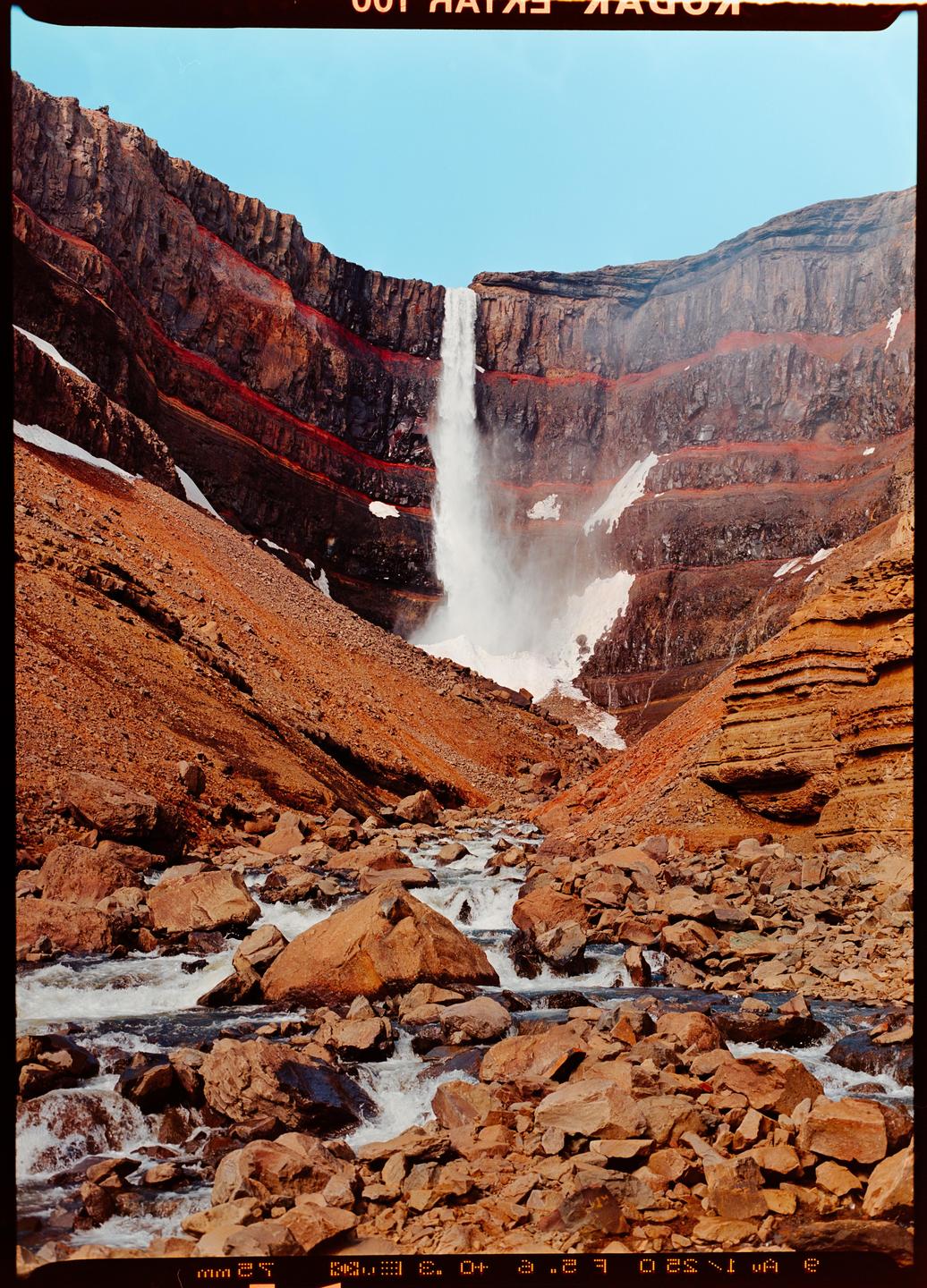 A powerful waterfall cascades down a red and brown rocky cliff into a riverbed filled with rocks and flowing water.