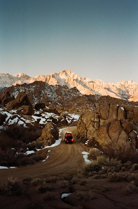 A red vehicle drives on a dirt road through a rocky, snow-dusted landscape with mountains in the background.