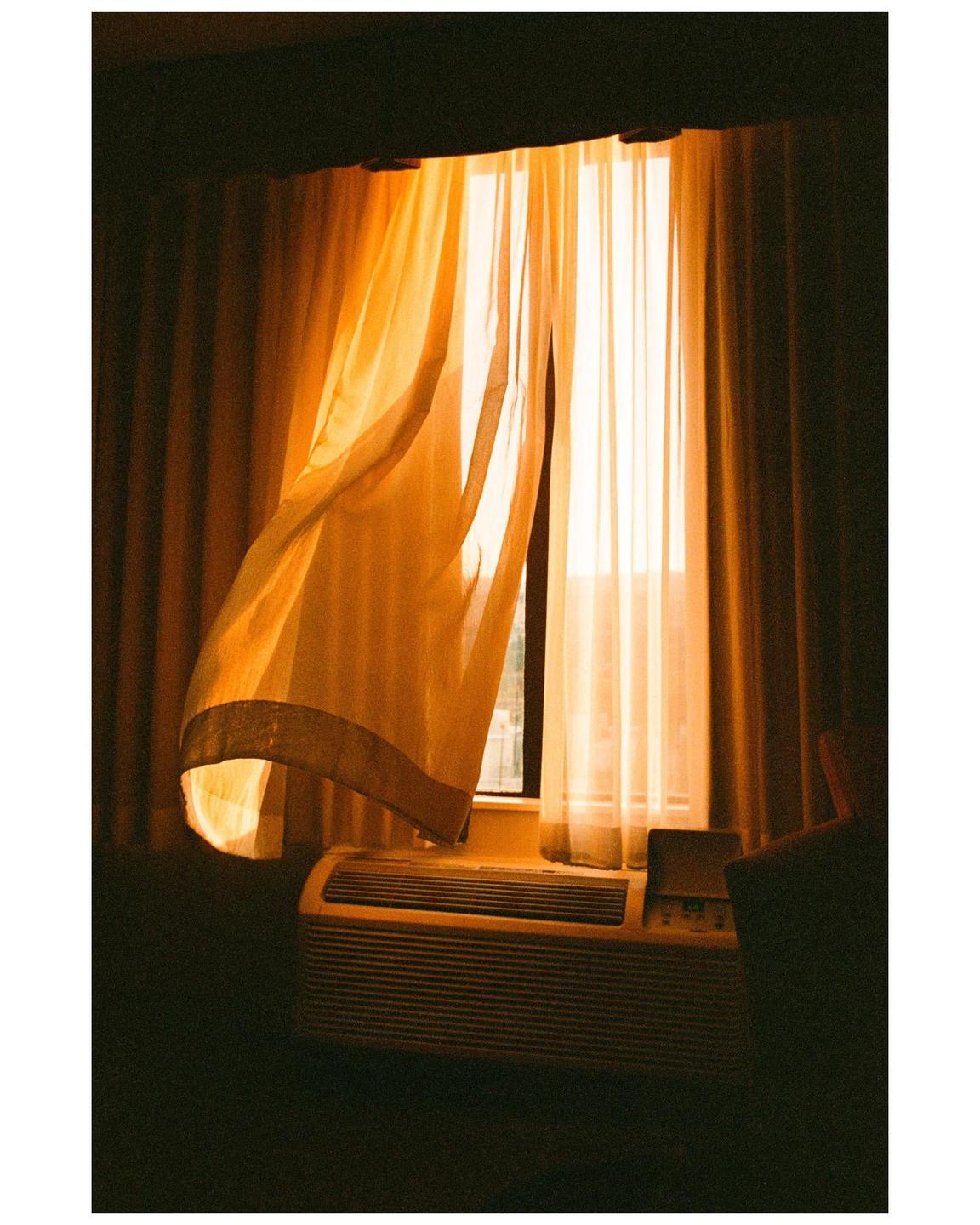 Sunlight streams through a window with sheer curtains and an air conditioner unit below.