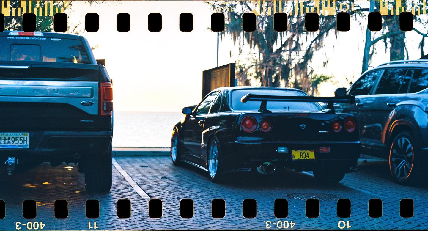 A black sports car with a large spoiler is parked between a pickup truck and another vehicle, with a body of water and trees in the background.