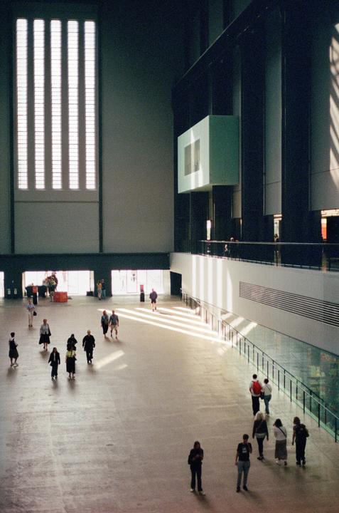People walk across a large, open hall with tall windows and a balcony.