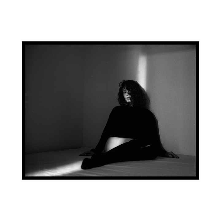 A woman with curly hair sits on a bed in a dimly lit room with a shaft of light illuminating the wall and bed.
