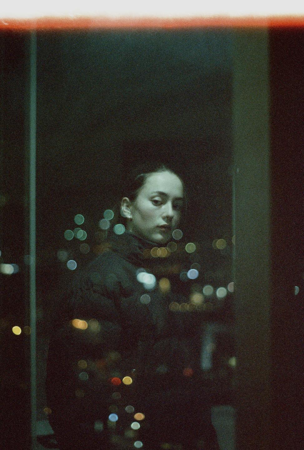 A woman stands in front of a window at night, with city lights visible as bokeh in the background.