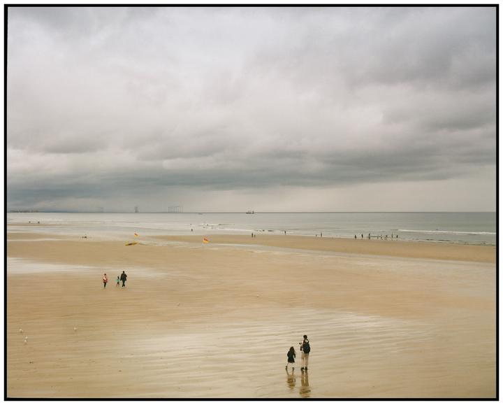 A wide sandy beach with people walking along the shore under a cloudy sky.