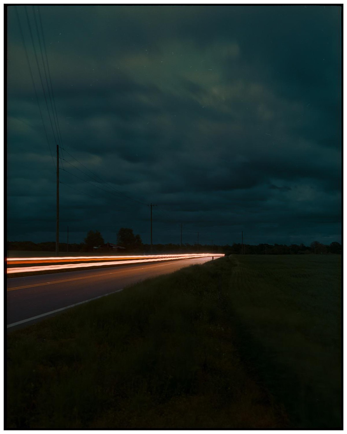 Light trails from a vehicle illuminate a rural road at night under a cloudy sky.