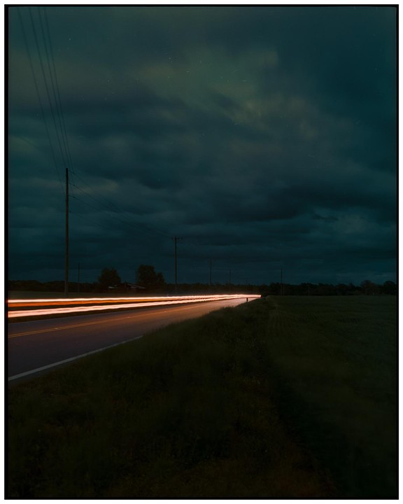 Light trails from a vehicle illuminate a rural road at night under a cloudy sky.