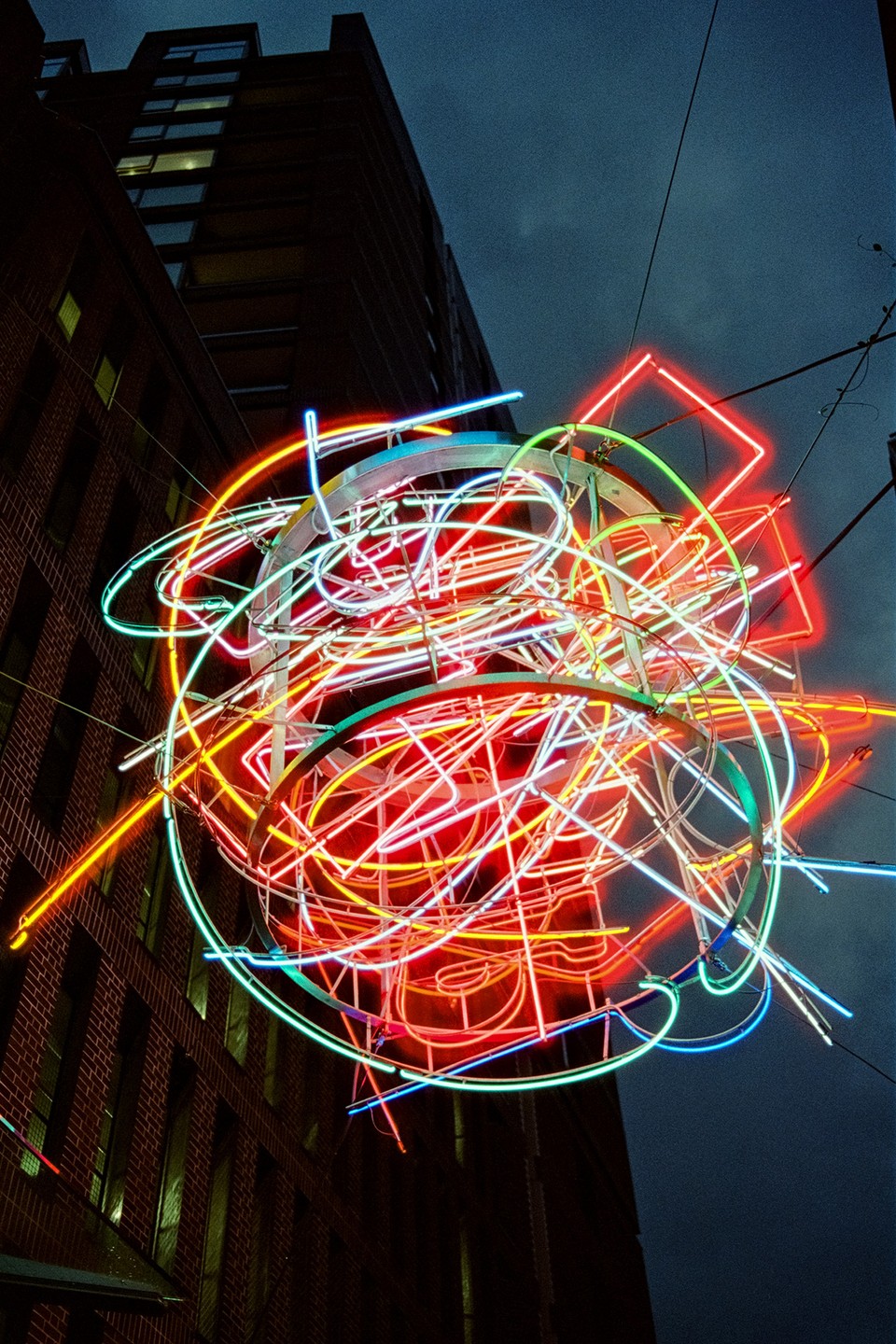 A colorful neon light sculpture hangs in front of a brick building at night.