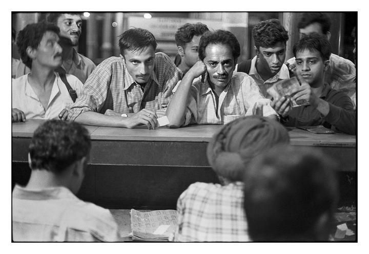 A group of men are gathered around a counter, some looking forward and one counting money.
